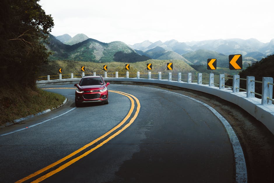 A red car drives along a winding mountain road with arrow signs and lush green scenery.
